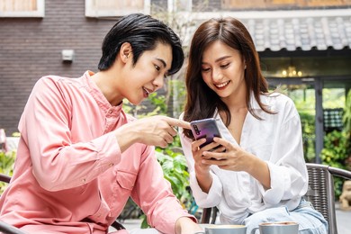 happy young asian woman sitting with boyfriend at cafe table, discussing over on smartphone, young couple looking at mobile phone shopping online and share information together in coffee shop