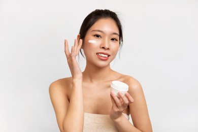 skin hydration and sun protection. portrait of cheerful asian lady holding jar and applying moisturizing creme or sunscreen lotion with spf on cheek and face, isolated on white studio background wall