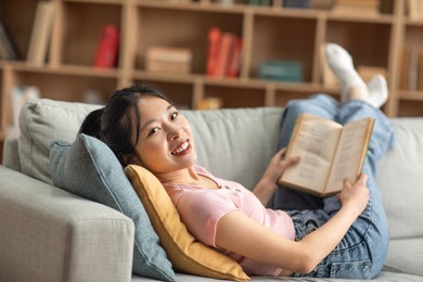 cheerful asian lady resting on couch in living room, reading book while spending time at home on weekend, looking at camera and smiling. hobby, domestic entertainment