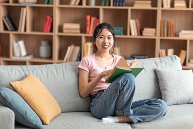 domestic life concept. positive asian lady holding paper book, sitting on comfy couch at home alone, reading interesting literature, enjoying story, free space