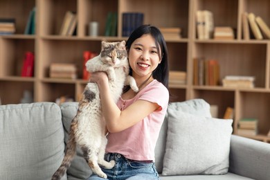 pretty korean lady posing with her beautiful cat, sitting on sofa in living room at home. happy chinese woman spending time with lovely pet, holding kitten and smiling at camera, copy space