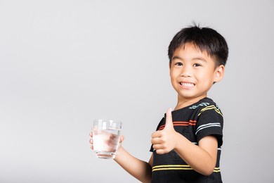 little cute kid boy 5-6 years old smile drinking fresh water from glass and show thumb up finger for good sign in studio shot isolated on white background, asian children preschool, daily life health