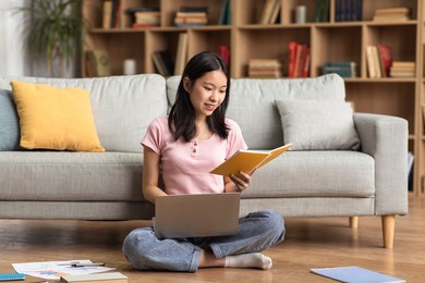 young korean lady studying online from home, reading book and using laptop pc, sitting on floor near sofa, free space. e-learning, web-based education concept