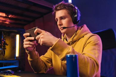 portrait of young guy pro gamer wearing headset streaming while playing mobile video game on his smartphone in neon coloured room at home. focused streamer participates in online e-sport tournament