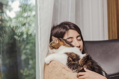 asian woman holding cat over her shoulder with clossed eyes and smile of happiness beside glass window and white curtain while cat looking out of window.