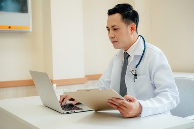 male doctor expert wearing white coat using laptop and reading clipboard report at work in hospital.