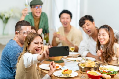 group of asian people enjoy dinner party eating food on dining table and using mobile phone taking selfie together at home. happy man and woman friends reunion meeting celebration on holiday vacation