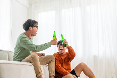group of asian man friends sitting on sofa watching soccer game competition on television with drinking beer together at home. happy male soccer fans cheering victory football team victory sport match