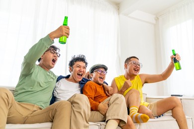 group of asian man friends sitting on sofa watching soccer game competition on television with drinking beer together at home. happy male soccer fans cheering victory football team victory sport match