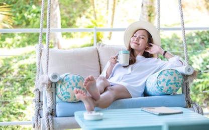 portrait image of a young woman with hat drinking coffee while relaxing and laying down on swing sofa 