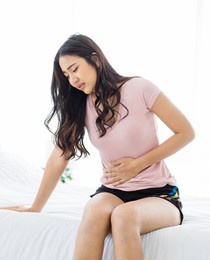 asian young stressed sick frowning face female model in casual outfit waking up in morning sitting on bed in bedroom holding hand on belly tummy having uncomfortable painful stomachache menstruation.