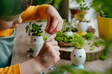 little girl decorating eggshells with grass with toy stickers eyes on the wooden table at home. creative fun diy idea for festive easter decoration. selective focus.