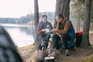 a married couple prepares food on a campfire on the shore of the lake with a tent. hiking camp with a bonfire in the forest. a man and a woman enjoy the silence by the fire near the tent