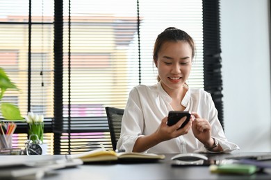 smiling asian young female office worker or businesswoman sitting at her office desk and using smartphone chatting with someone.
