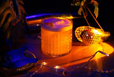 a closeup of a gin fizz cocktail, lemon twist garnish, and bar equipment under neon light
