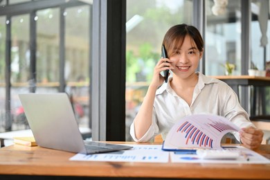 professional asian female financial consultant talking on the phone with her client while reviewing financial report in the office.