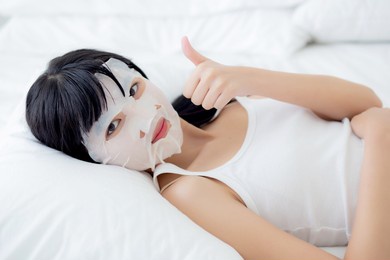 beautiful young asian woman with sheet facial mask and thumbs up on bed at bedroom, beauty girl lying applying cosmetic and makeup with skin care face for wrinkle, skincare treatment and health.