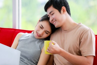 portrait shot of an adorable young asian lover couple with cute smiling sitting on the sofa and watching movies on laptop together. the wife sitting beside her husband and snuggle him with love.