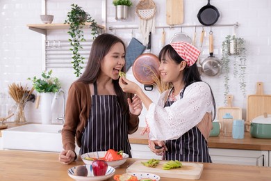 cheerful asian girl feeding her female friend with fruit at home kitchen.smiling beautiful women preparing salad together.cute sister preparing healthy food for family party fruit,salad,happiness.