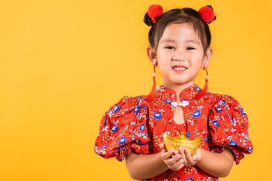 chinese new year. happy asian chinese little girl smile wearing red cheongsam holding gold ingot, portrait children in traditional dress hold golden bar, studio short isolated on yellow background