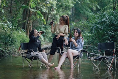 group of asian girls enjoying a day at the  during holiday camping . sitting on a camping chair dip them legs in the river and drinking cold beer from the bottle . 