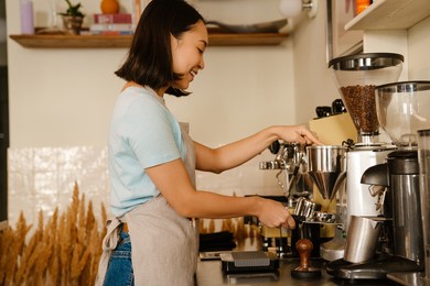 young asian barista woman wearing apron smiling while making coffee in cafe