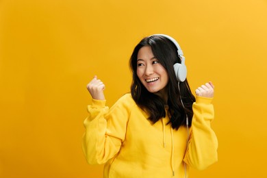 cheerful happy asian student young woman in yellow hoodie sweatshirt enjoy good mood today posing isolated on over orange studio background. cool offer. perfect playlist sound app concept