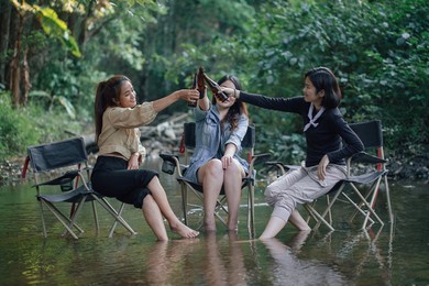 group of asian girls enjoying a day at the  during holiday camping . sitting on a camping chair dip them legs in the river and drinking cold beer from the bottle.