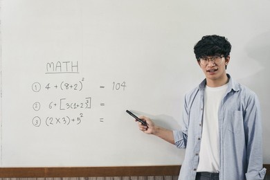 asian male teacher is teaching students at the classroom while pointing at numbers on the white board.