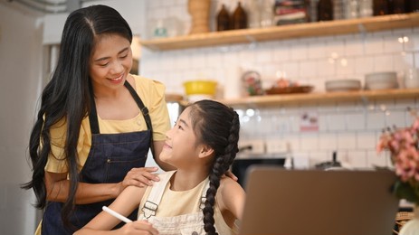 smiling asian mother helping her little daughter with distance learning virtual distance online class on laptop computer