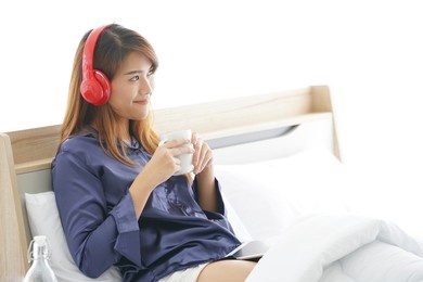 happy young asian woman sitting in bed and holding white cup and listening with red headphones