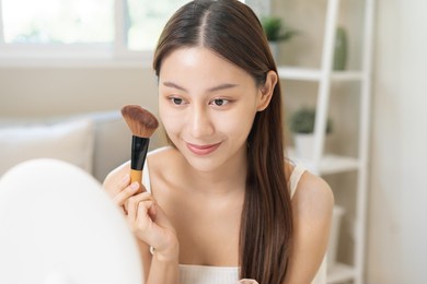 happy beauty blogger concept, cute asian young woman, girl smile, make up face by applying brush blush powder on her cheek, looking at the mirror. people look with natural fashion style.