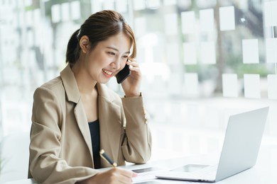 overjoyed asian female laughing telling funny story by phone to friend colleague sitting by office computer screen. young woman enjoying conversation by cell discussing good positive news.