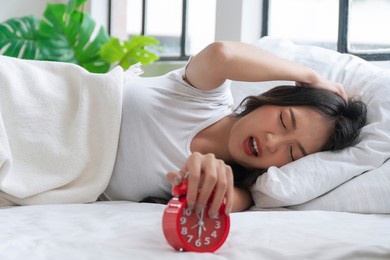 upset asian young woman displeased stopping ringing red alarm clock on white bed in morning. angry unhappy young girl sleeping with white pillow holding hand bell snooze alarm clock laziness at home.
