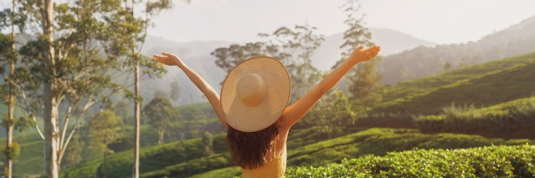 carefree woman traveler with raised hands standing against natural background tea plantations landscape. back view of young brunette model in straw hat looking away in front of perfect natural