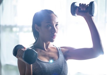 asian young woman doing dumbbell workout at home, working on arms strength, athletic female lifting blue fitness dumbells up over living room interior