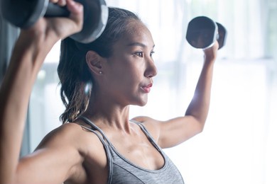 asian young woman doing dumbbell workout at home, working on arms strength, athletic female lifting blue fitness dumbells up over living room interior