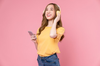 cheerful young asian woman in headphones listening to music and enjoy favourite playlist application on smartphone with dancing on pink background.