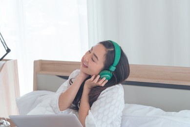 young asian woman wear green headphones relaxing and listening the music while using laptop computer on the bed, girl enjoys audio sound, relaxing