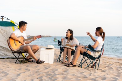 happy friend have fun playing guitar and clap in camp they smiling together in holiday on sand beach near camping tent vacation time at sunset, young asian group woman and man in summer travel outdoor