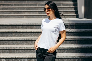 stylish brunette asian girl wearing white t-shirt and glasses posing against street , urban clothing style. street photography