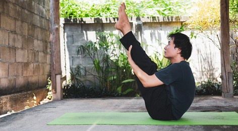 healthy living concept of young asian man practicing yoga asana paripurna navasana pose or full boat pose, work out and poses on a green yoga mat. outside exercise in the garden. healthy lifestyle.