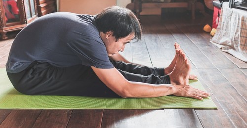 young asian man practicing yoga in a wooden room sitting in seated forward bend exercise, paschimottanasana pose on green mat yoga at wooden home. healthy living.