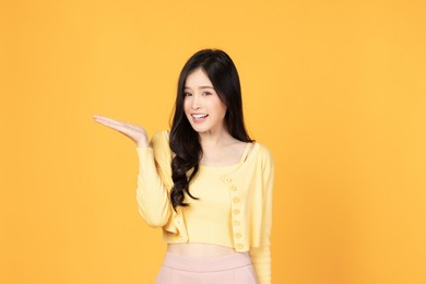 beautiful asian woman wearing casual clothes smile and pointing finger to an empty palm isolated on yellow background studio portrait.