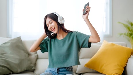 young asian woman listening to music on couch in living room at home. happy asia female using mobile smartphone, wearing headset and sitting on sofa