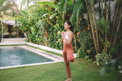 indonesian girl practicing yoga in tree pose outdoor. concept of harmony and mental health. young smiling athletic woman wearing sportswear standing barefoot on green grass near swimming pool in yard