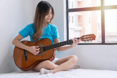 cheerful asian woman casual playing acoustic guitar sitting on white bed beside window at apartment. enjoy player young girl play acoustic guitar relax hobby at home.lifestyle instrument music concept