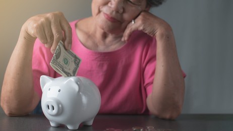 finance and savings concept, old woman in pink shirt is putting banknotes in a white piggy bank lying on the table.