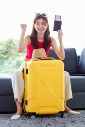 asian young happy cheerful female traveler in casual summer outfit with sunglasses sitting smiling on cozy sofa in living room holding passport and boarding pass ticket with yellow trolley luggage.