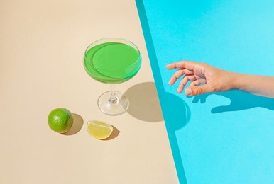 creative composition with woman hand reaching for cocktail and sliced lime against duo tone sandy and blue background. sunny summer scene. minimal pool party concept.